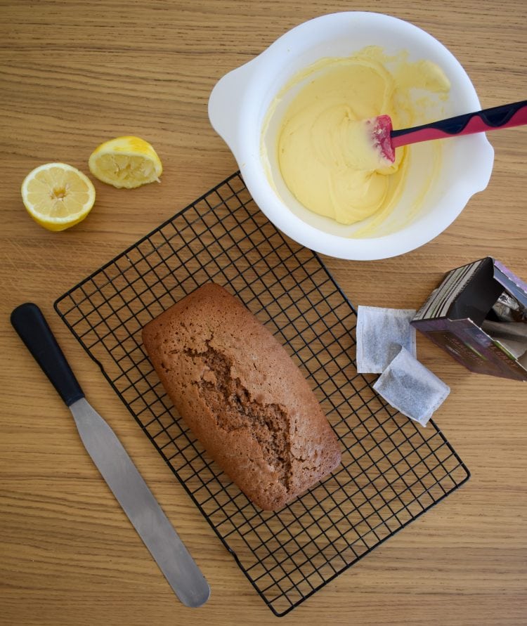 Un-topped coffee loaf cake on wire rack. Palette knife, teabags and sliced lemon to the side. Lemon buttercream in mixing bowl at the top of shot. Top view.
