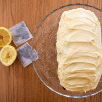 Earl Grey Loaf Cake with Lemon Buttercream on glass cake stand, sliced lemons and teabags to left of shot