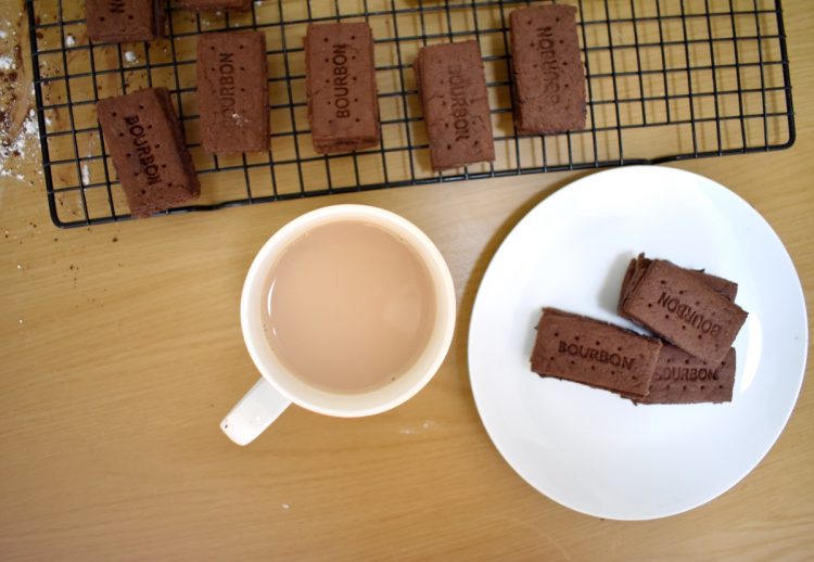 Top view, cup of tea next to Bourbon biscuits on plate, other biscuits cooling on wire rack