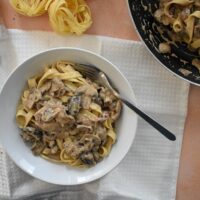 Top view of served mushroom stroganoff with pasta in bowl, frying pan leaving shot, salt, pepper and pasta on table