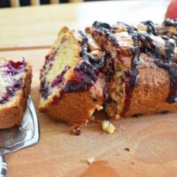 Apple blackberry loaf cake on chopping board, one slice flat, another leaning against the cake, apples in background