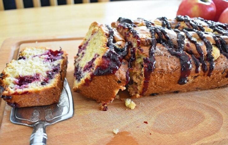 Apple blackberry loaf cake on chopping board, one slice flat, another leaning against the cake, apples in background