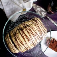 Bailey's Cake on glass cake stand, surrounded by bottle, slice of cake on a plate and white towel, shot from above