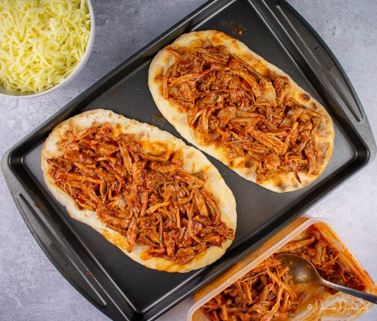 2 flatbreads with pork on baking tray, bowl of cheese and pulled pork to sides.