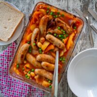 Sausage and tomato bake in glass cooking dish. Serving bowls and bread to the sides.