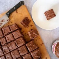 top view, nutella traybake on chopping board with knife. Small square of cake on white plate. Open jar of nutella also in shot.