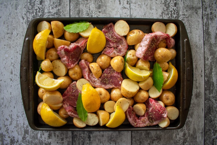 Uncooked lamb chop traybake with potatoes, lemons and bay leaves on baking tray.