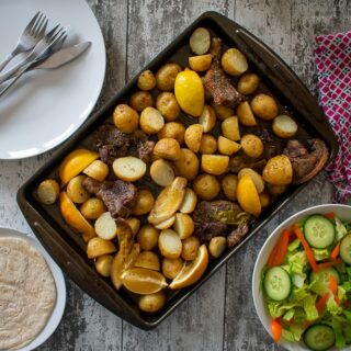 cooked greek lamb traybake on baking sheet. Salad, bread and empty plates surround.