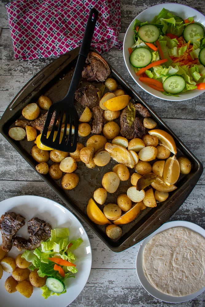 baking sheet with lamb and potatoes in the middle, served portion and salad bowl to the sides.