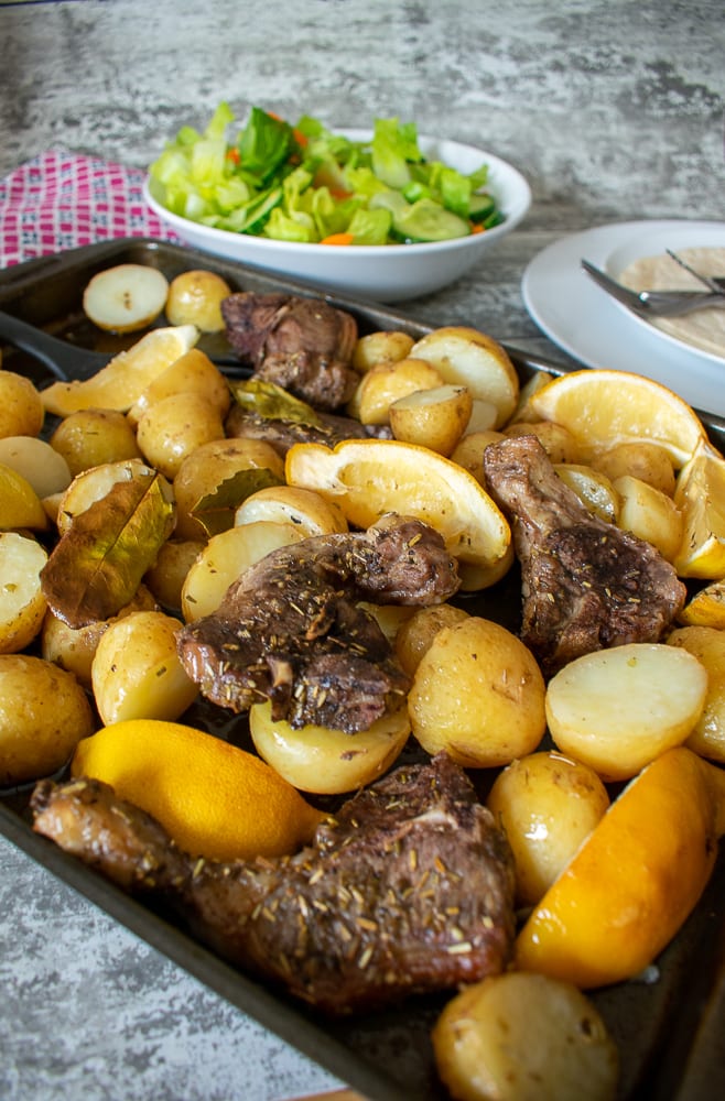 lamb chops, lemons and potatoes on baking tray, salad in bowl in the background.