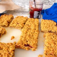 3/4 shot of jam flapjack on chopping board, jar of jam with spoon in background.