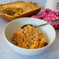 served portion of crumble in small white bowl, jug of custard and baked crumble in the background.