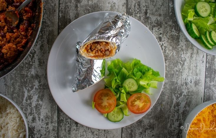 sausage burrito wrapped in foil, on a plate with side salad.