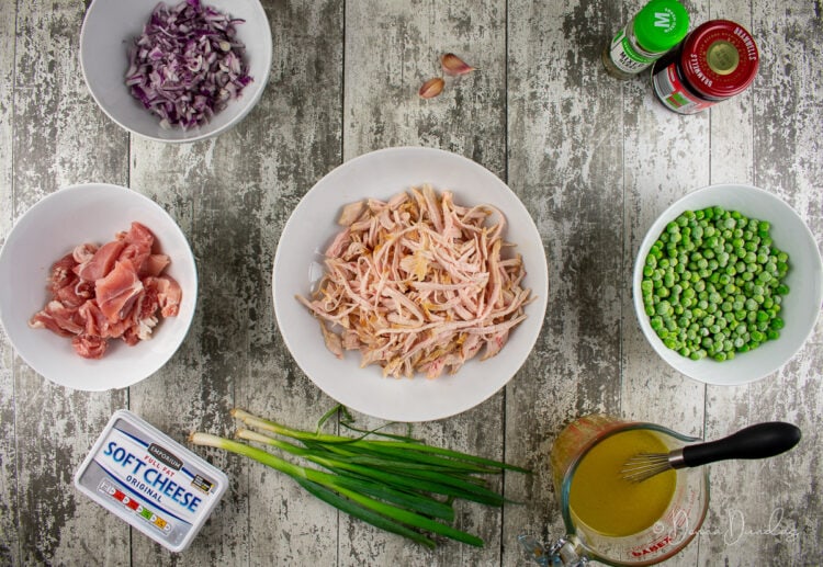 ingredients for leftover turkey pasta. White bowls. Shredded turkey, diced red onion, chopped bacon, soft cheese, spring onion, jug of stock, frozen peas, jar of cranberry sauce, mixed herbs, cloves of garlic.