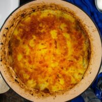 leftover lamb shepherd pie in cast iron dish on blue table cloth. Empty bowls, plate with bread and serving spoon to the side. Close up.