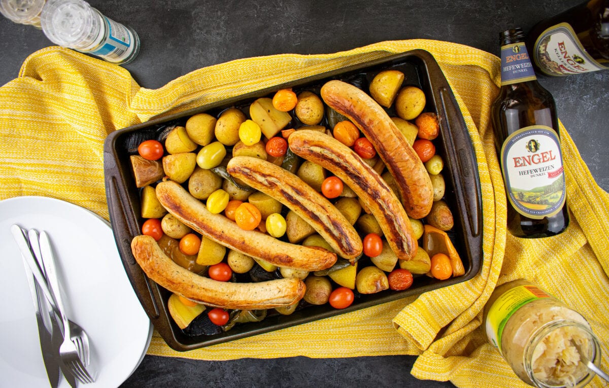 Baking tray on yellow cloth with large bratwurst, potatoes, peppers and tomatoes. Bottles of beer, sauerkraut and empty white plate to the sides.