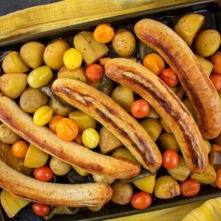 Baking tray on yellow cloth with large bratwurst, potatoes, peppers and tomatoes.