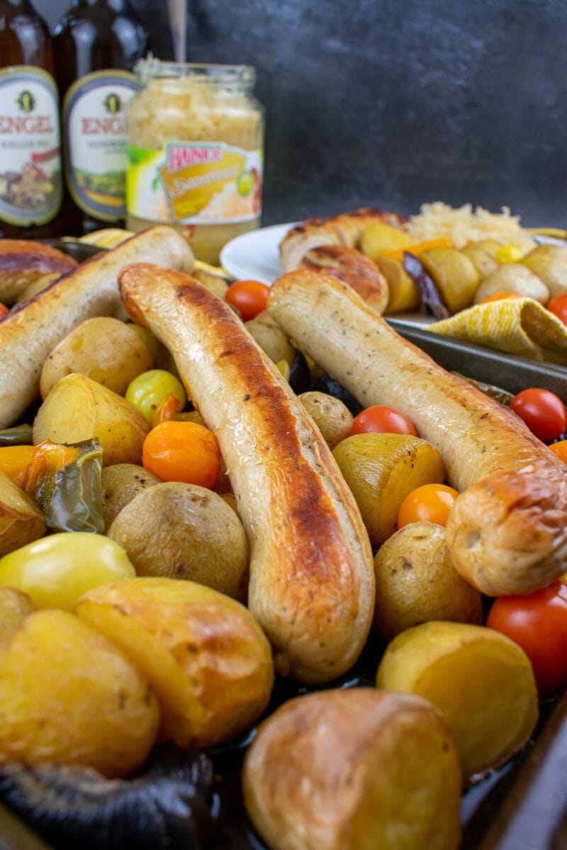 bratwurst, potatoes, tomatoes and peppers on baking tray. Jar of sauerkraut, bottles of beer and served plate of food in the background.