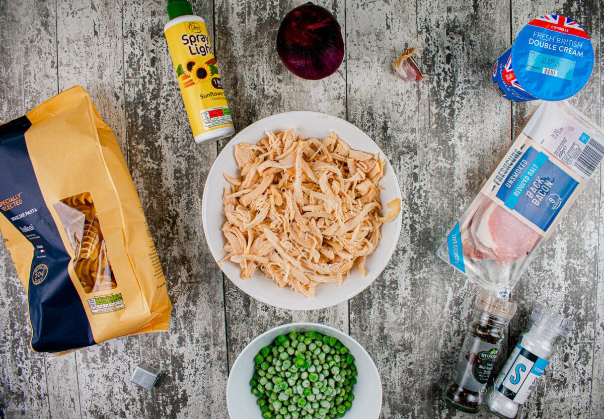 Ingredients for leftover chicken pasta on table - bag of pasta, bowl of shredded chicken, bowl of peas, stock cube, salt and pepper, pack of bacon, double cream, garlic cloves, red onion and cooking spray.