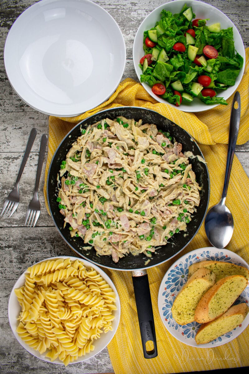 leftover chicken pasta with peas in black frying pan in middle of table on yellow cloth. Empty bowls, bowl of salad, plate with garlic bread and bowl with fusilloni pasta around. Flatlay.