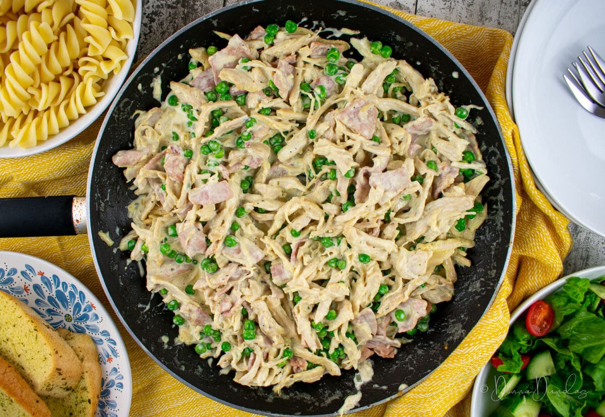 close up of chicken pasta in black frying pan from above. Salad bowl, garlic bread on plate and bowl of pasta to the sides, just in shot.