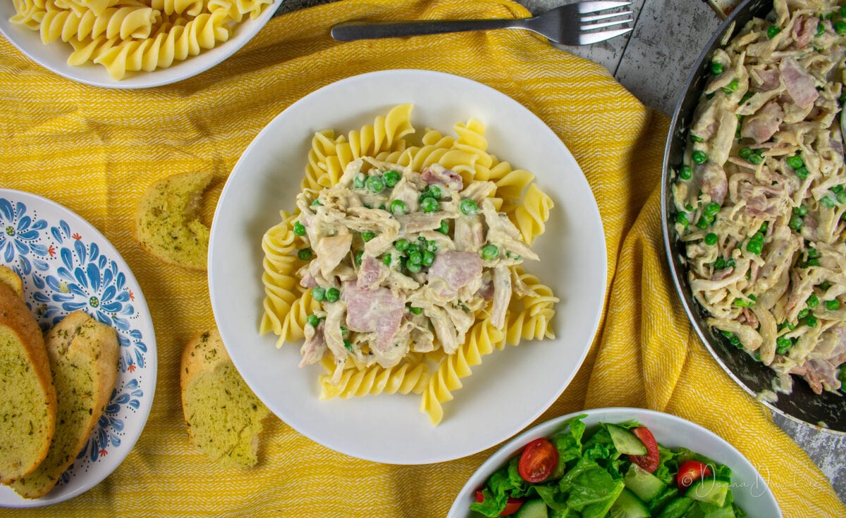 served portion of leftover chicken pasta in white pasta bowl on yellow cloth. Frying pan, plate with garlic bread, pasta in bowl and salad bowl all leaving shot in corners.