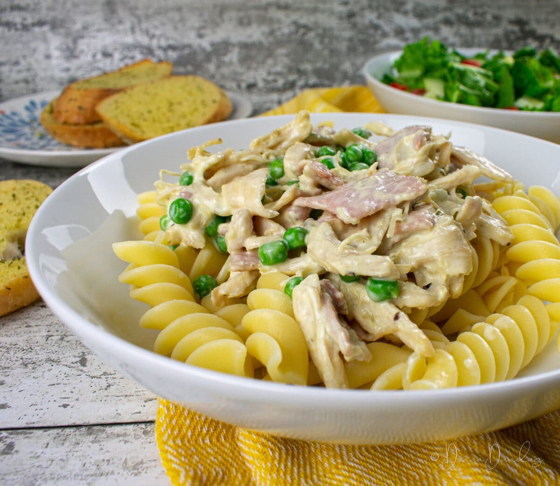 3/4 angle shot of served chicken, bacon, peas, cream sauce and pasta. Bowl of salad and plate of garlic bread blurred in background.