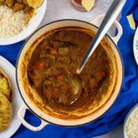 Lamb curry in cast-iron dish with serving spoon. Curry sauce brown/red, chunks of tomato and lamb visible. Plate with naan bread, poppadoms, bowl of rice and empty bowl leaving shot in the corners.