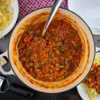 cooked ragu in cast iron dish, bowl of pasta to the side, served portion with tagliatelle in white pasta bowl at the bottom.