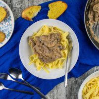 top view of served portion of leftover beef stroganoff with tagliatelle in pasta bowl. Frying pan, bowl of pasta and plate of garlic bread leaving shots to the sides.