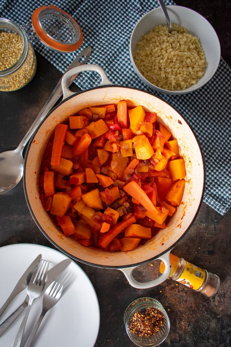 veggie tagine in cast-iron dish on table. Served portion on white plate to the side.