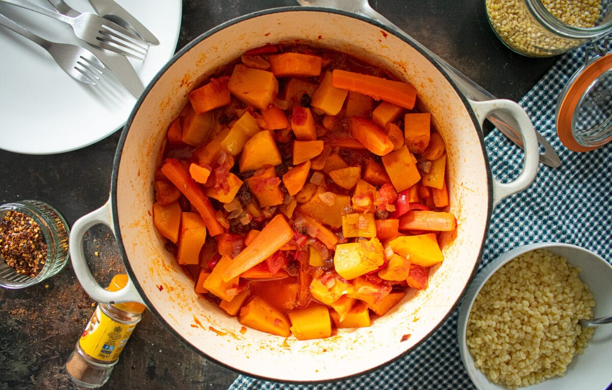 veggie tagine in cast-iron dish on table. Served portion on white plate to the side.