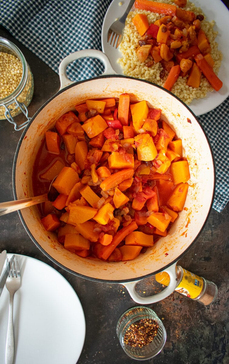 veggie tagine in cast-iron dish on table. Served portion on white plate to the side.