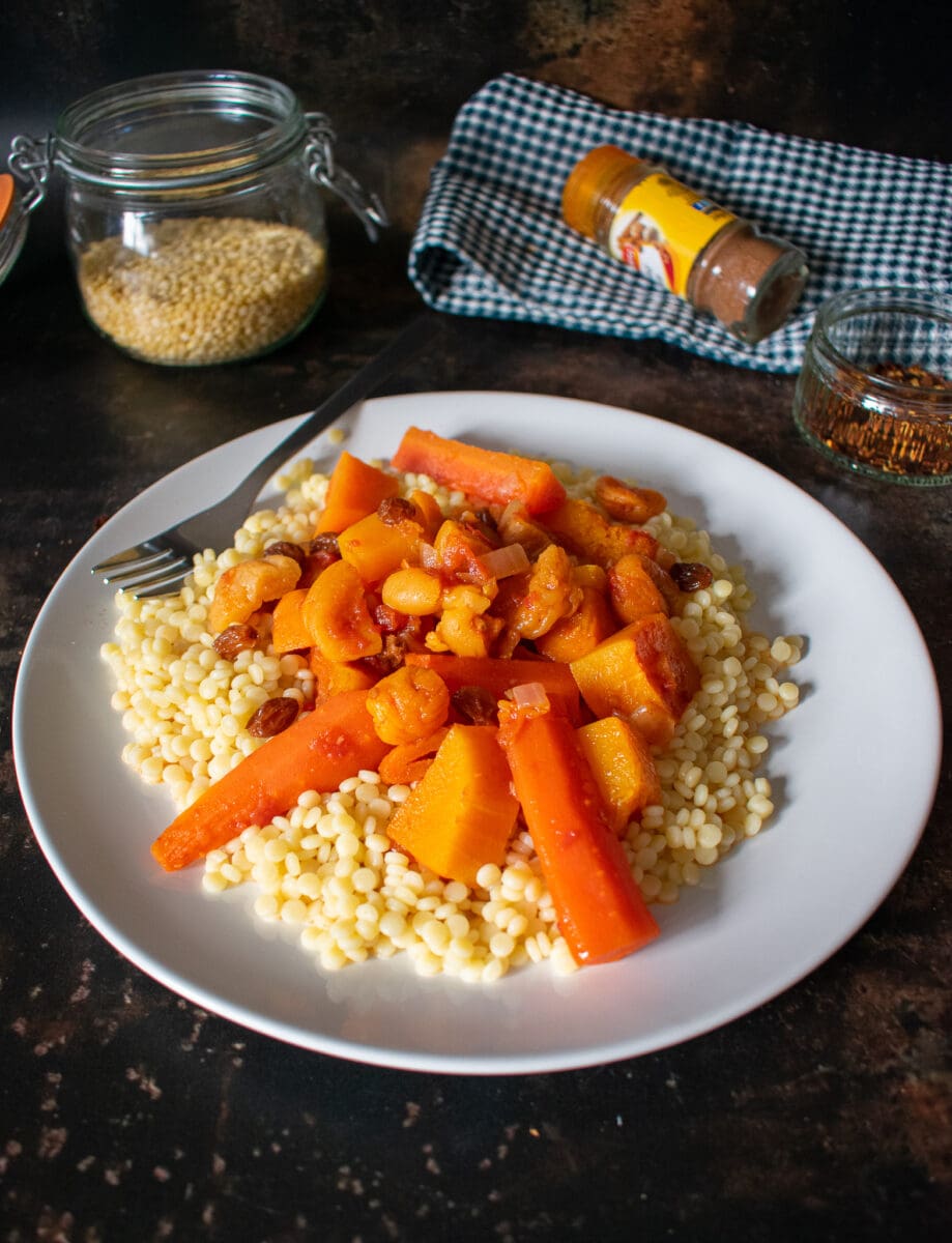 side view close of of tagine on plate with couscous, bowl of couscous, and spices in the background on tea towel.
