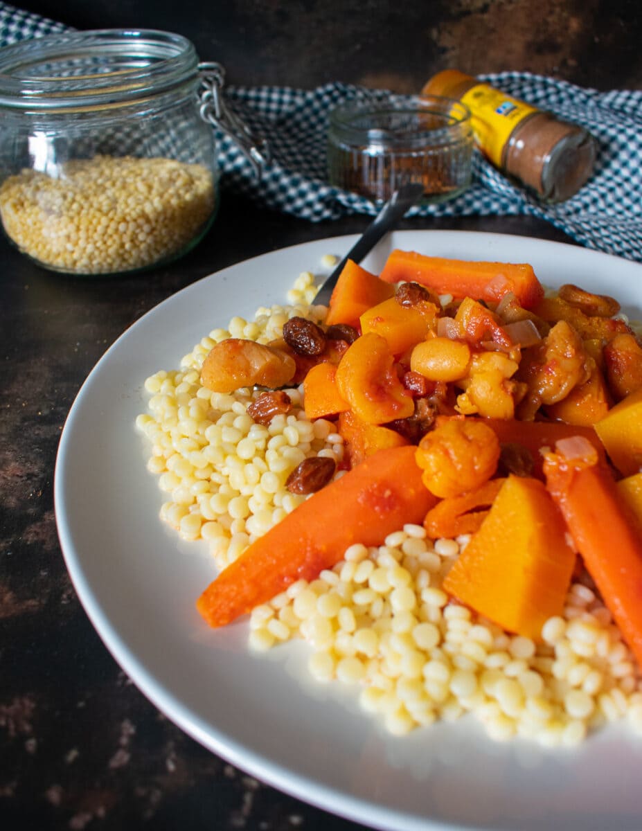 side view close of of tagine on plate with couscous, bowl of couscous, and spices in the background on tea towel.