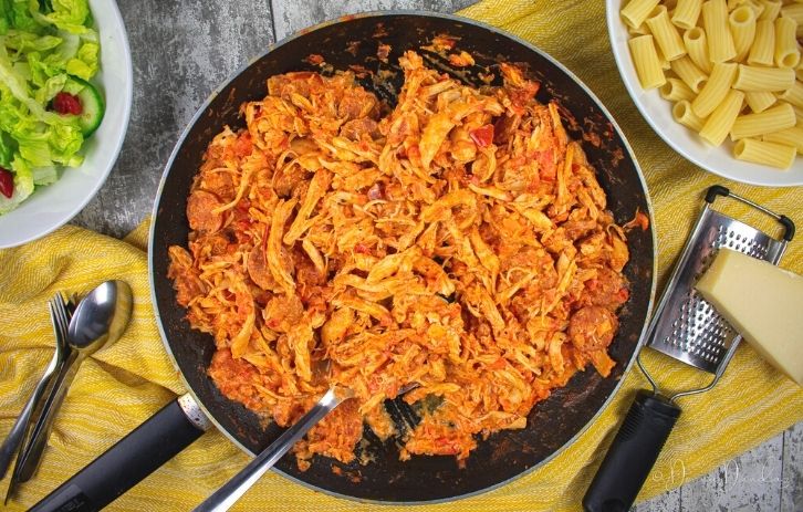 Chicken and chorizo pasta in large frying pan. Bowl of pasta, salad and block of cheese with grater also on the table.