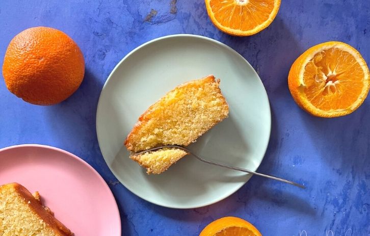 served slice of orange drizzle cake on green plate with fork. Oranges round the sides, leaving frame.