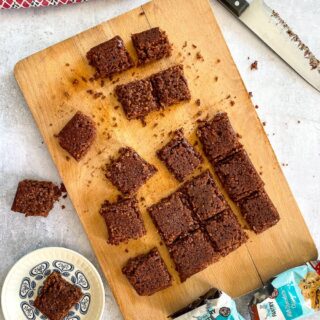 top view of squares of chocolate flapjacks on chopping board. One square on small blue and white plate to the side. Also in shot - sharp knife, red towel and empty chocolate chip packets.