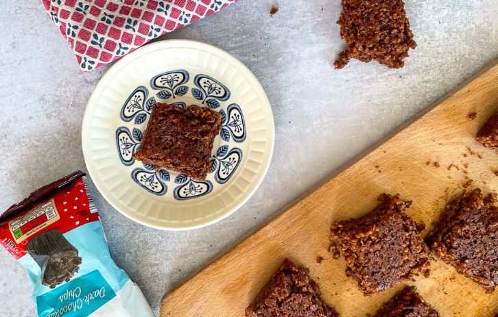 top view of squares of chocolate flapjacks on chopping board. One square on small blue and white plate to the side. Also in shot - sharp knife, red towel and empty chocolate chip packets.