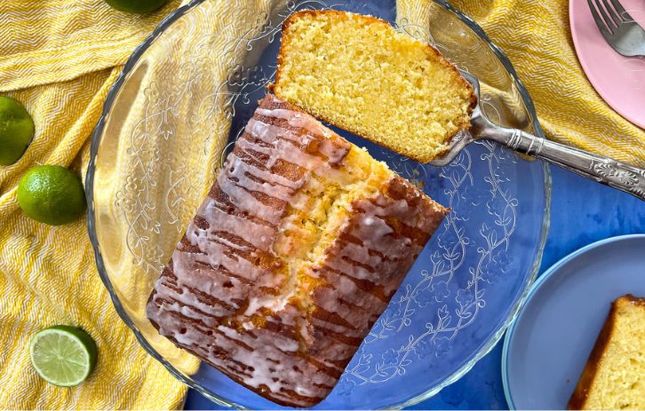 Lime drizzle cake on glass cake stand. One slice laying flat on a silver antique cake slice. Cut limes and served portions of cake on plates to the side.