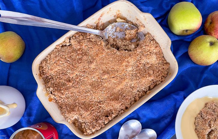Apple and caramel crumble in baking dish with silver serving spoon. Served portion in bowl above, apples on side.