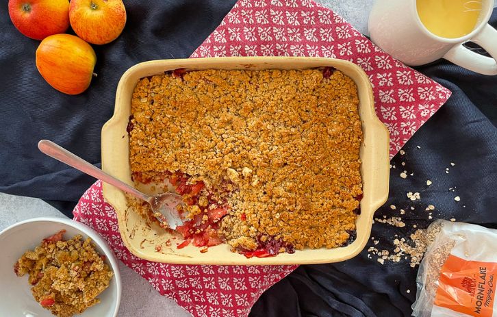 apple and raspberry crumble in baking dish, served portion in bowl to the side. Dish surrounded by a jug of custard, 3 red apples and an open bag on oats.