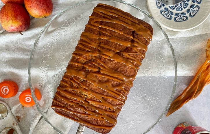 Apple and cinnamon loaf cake on glass cake stand. Slice of cake on plate to side. Surrounded by tin of caramel, apples and spice jars.