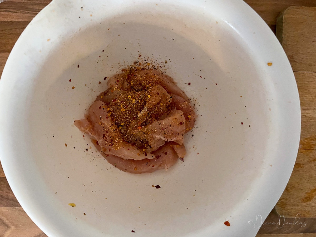 uncooked chicken with oil and peri peri seasoning in white mixing bowl.