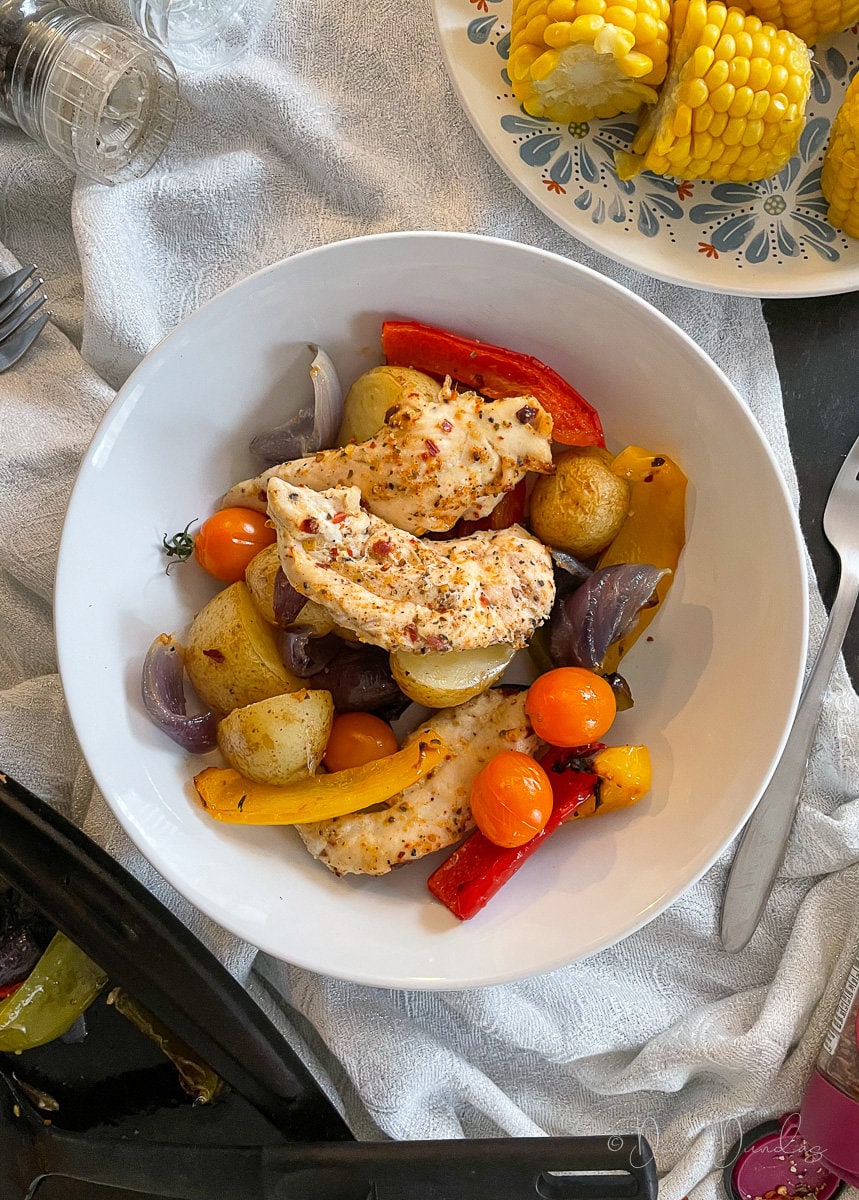 top view of bowl with peppers, potatoes, onions, tomatoes and chicken, plate of corn in the background.