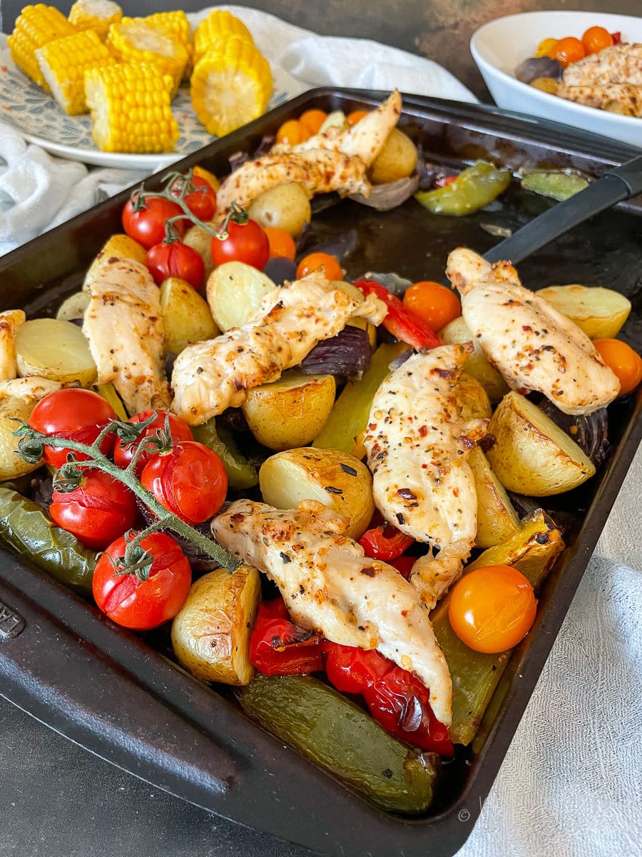 side view of cooked peri peri traybake ingredients in baking tray. Served portion and plate of corn in the background.