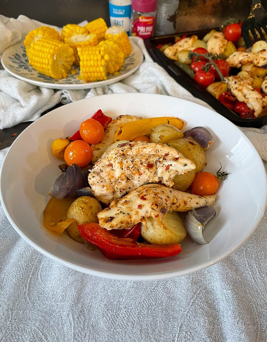 side view of served portion of peri peri traybaek in white bowl. Plate of corn, and baking tray in the background.