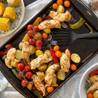 baking tray wiht chicken fillets, potatoes, peppers, onion and tomatoes. Spatula in tray, served portion to the side in pasta bowl. Side plate with corn on the cob.