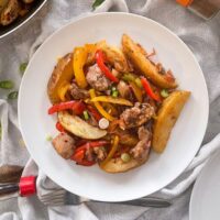 Salt and pepper chicken with peppers and potatoes served on white plate. Frying pan leaving shot, soy sauce, salt and pepper and empty plate also on table.