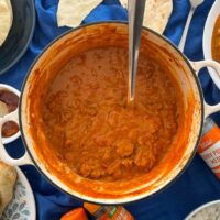 lamb tikka masala in cast iron dish with serving spoon. Served potion, naan bread, poppadoms, and bowl of rice also on table on blue cloth.